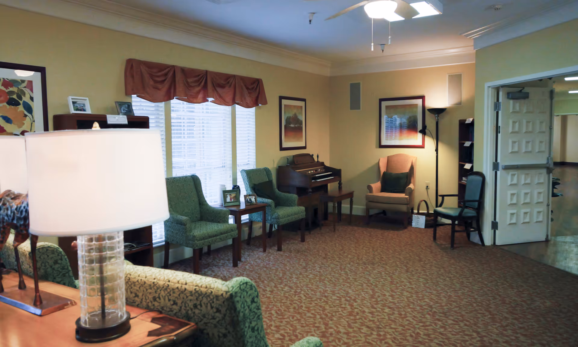 A cozy living room area with patterned carpet, several upholstered chairs, a small wooden table with framed photos, a lamp with a clear textured base, a piano against the wall, framed artwork, and a ceiling fan with lights. There is a doorway leading to a hallway on the right side.