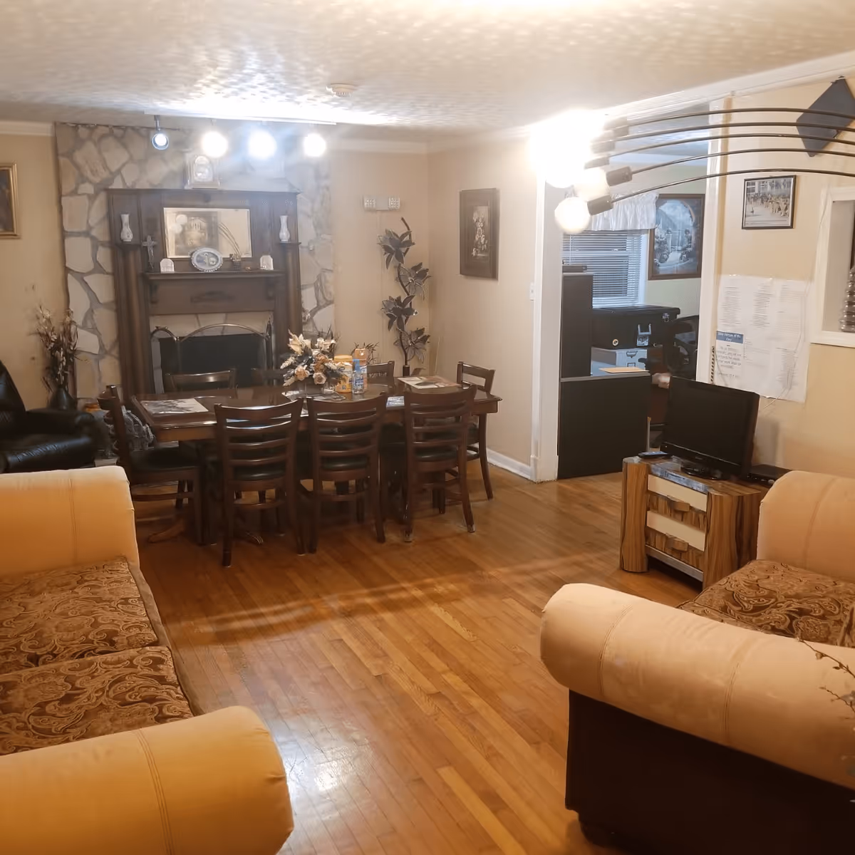 A cozy living and dining area with wooden floors, a dining table with eight chairs, a stone fireplace with a wooden mantle, two beige sofas with patterned cushions, a small TV on a wooden stand, and various wall decorations and lighting fixtures.