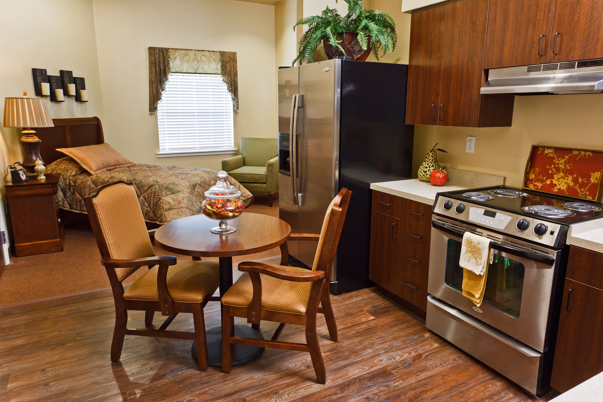 Studio-style senior living interior with a bed, small round dining table and chairs, and a kitchenette with stainless steel appliances.