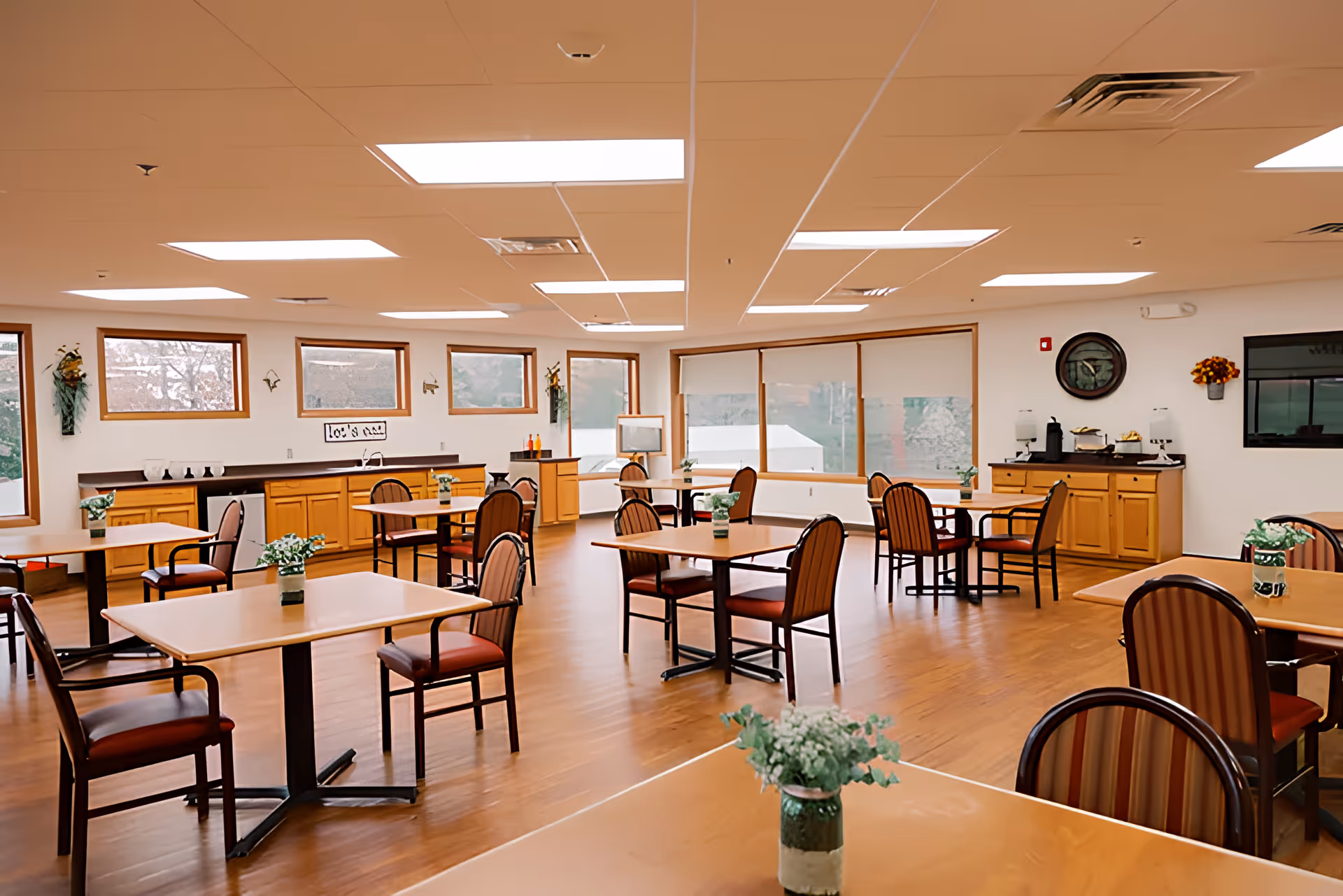 A bright and spacious dining room with multiple square tables and chairs arranged neatly. Each table has a small green plant centerpiece. The room has large windows allowing natural light to fill the space, wooden flooring, and light-colored walls. There are cabinets and countertops along the walls, with some decorative items and a coffee station visible.