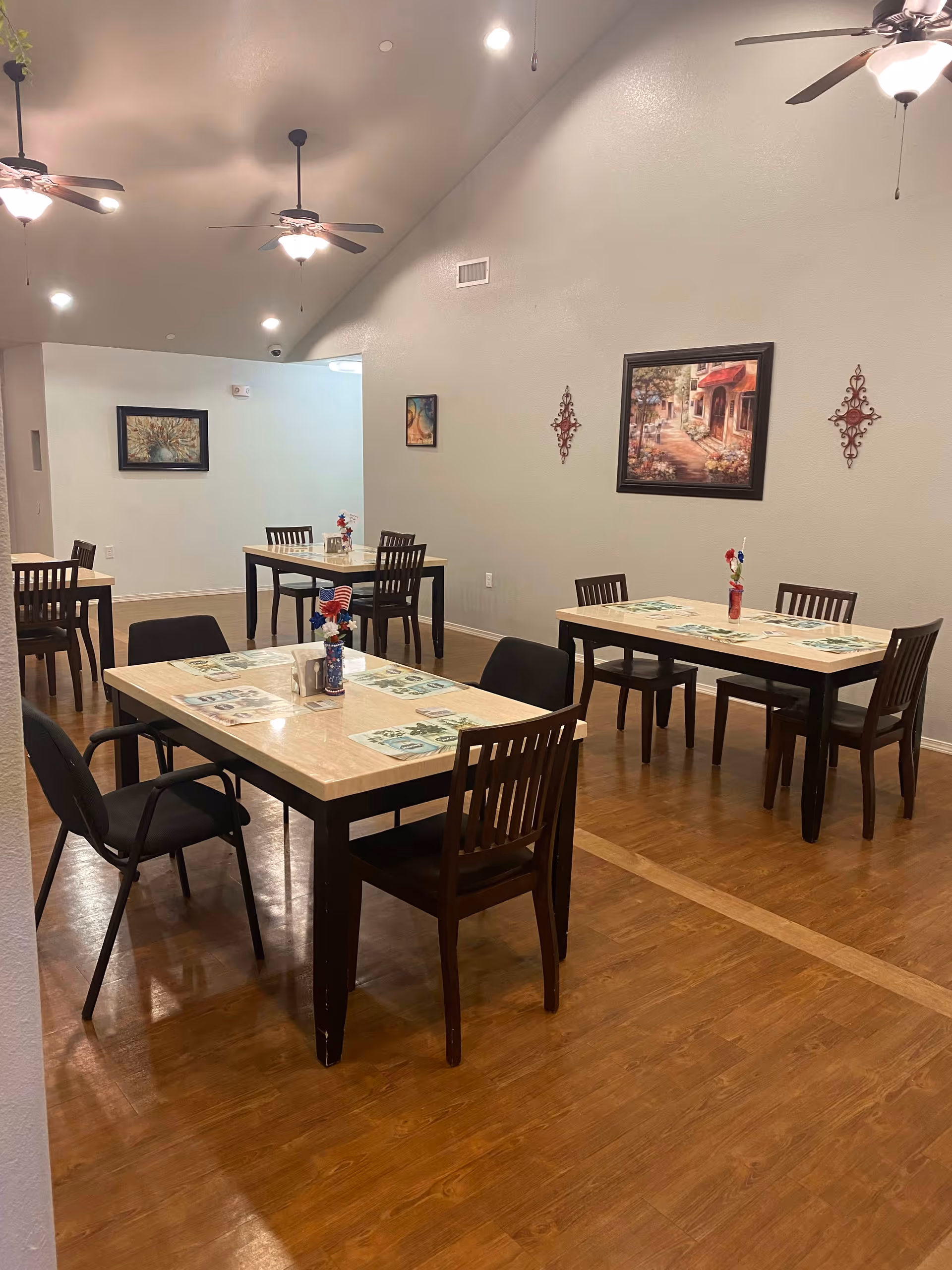 Interior view of a dining room with several tables and chairs arranged neatly. Each table has placemats and a small vase with decorative flowers. The room has wooden flooring, ceiling fans with lights, and artwork hanging on the walls.