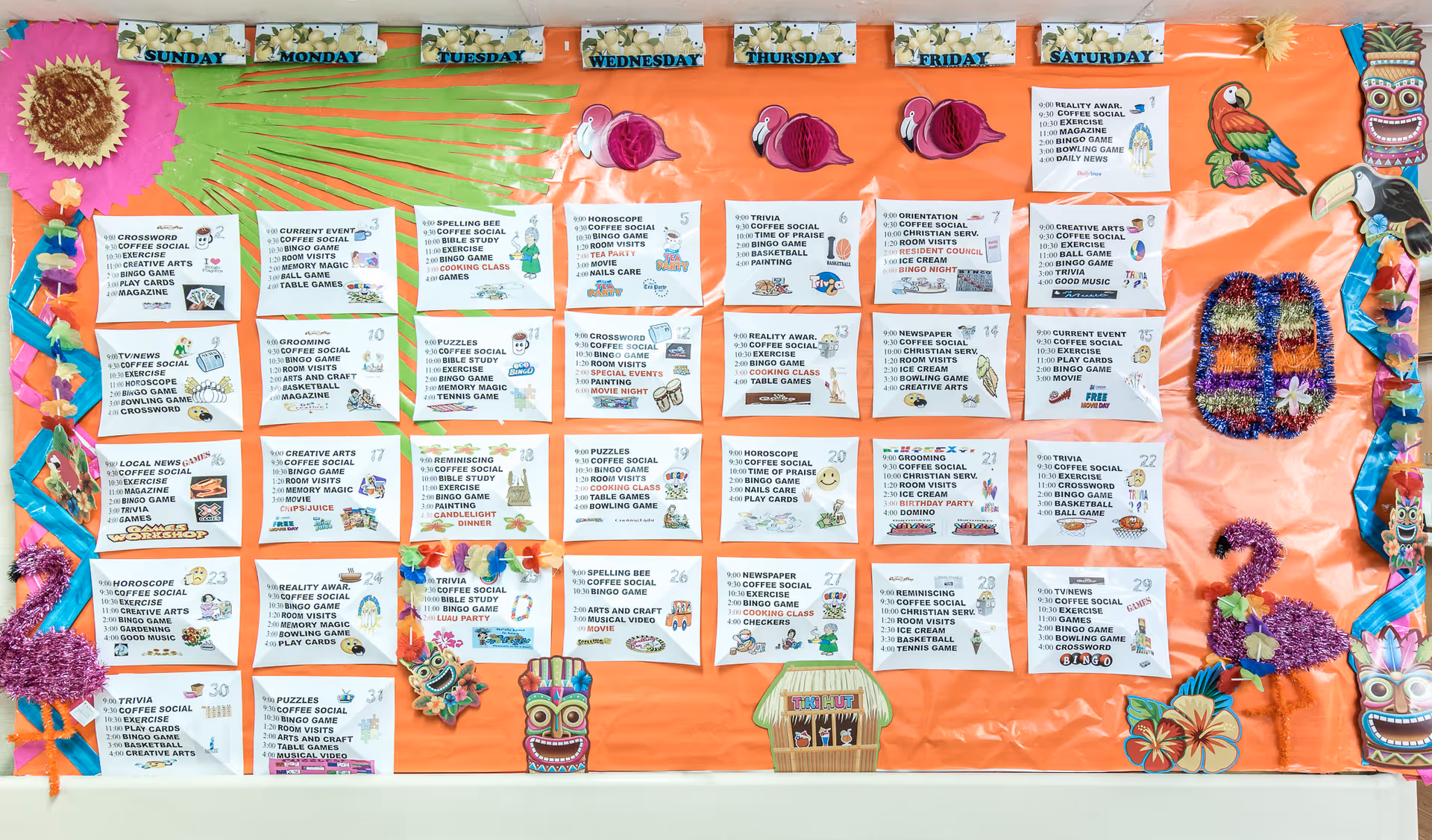 A colorful monthly activity calendar displayed on an orange bulletin board decorated with tropical-themed items such as flamingos, tiki masks, parrots, flowers, and palm leaves. The calendar lists daily activities including crossword, coffee social, exercise, bingo, games, and various arts and crafts events. Days of the week are labeled at the top with decorative headers.