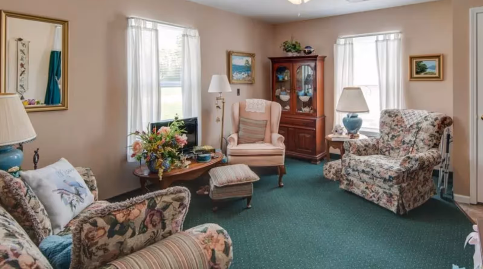 A cozy living room with floral patterned armchairs and sofa, a pink armchair with a striped cushion, a wooden coffee table with a flower arrangement, a wooden cabinet with glass doors, two windows with white curtains, and two table lamps with blue bases.