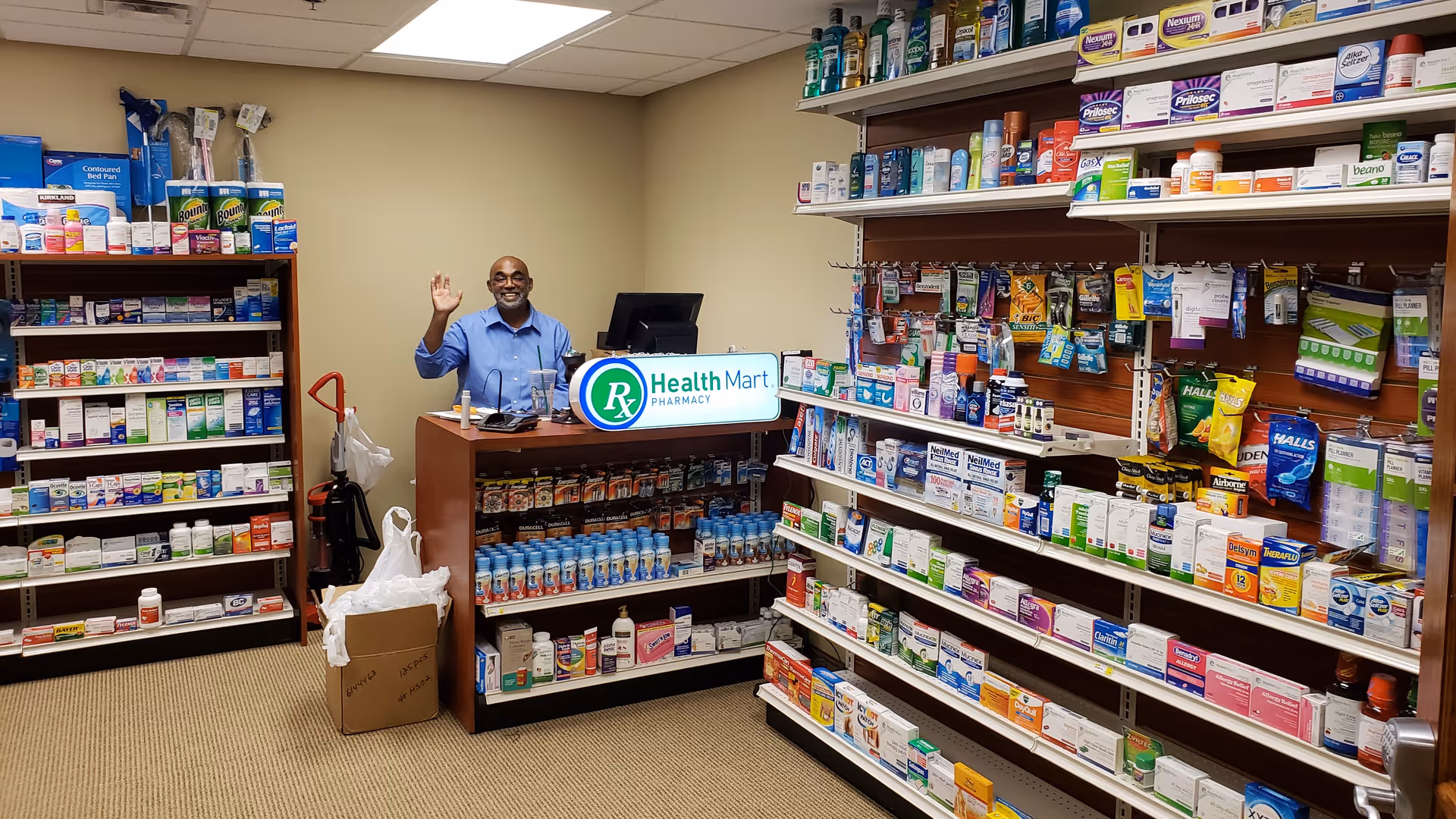 A man standing behind a pharmacy counter inside a room with shelves stocked with various medications and health products. The man is smiling and waving. The counter has a sign that reads 'Health Mart Pharmacy'.