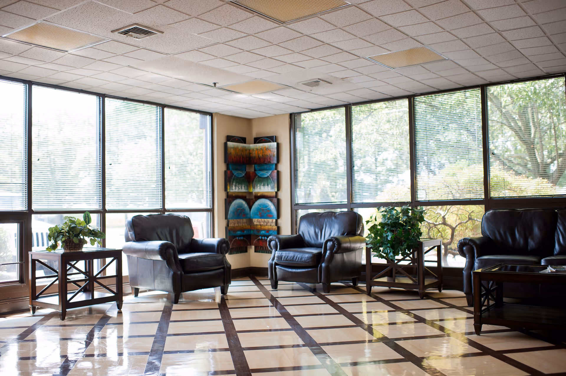 A bright and spacious living room area with large windows covered by blinds, three black leather armchairs, two wooden side tables with green plants, and a colorful abstract wall art piece in the corner.