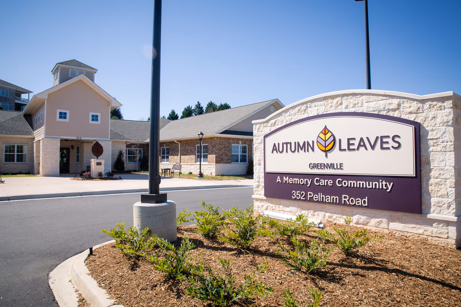 Exterior view of a memory care community building with a large stone sign in the foreground that reads 'Autumn Leaves Greenville, A Memory Care Community, 352 Pelham Road'. The building has a beige and brick facade with a peaked roof and several windows. There are small bushes and a lamp post near the sign.