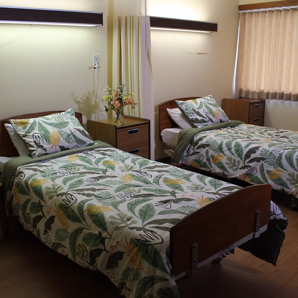 A room with two single beds featuring matching green and yellow leaf-patterned bedding. Each bed has a wooden headboard and footboard. Between the beds is a wooden nightstand with a vase of flowers. The room has beige walls, a window with beige curtains, and overhead lights above each bed.