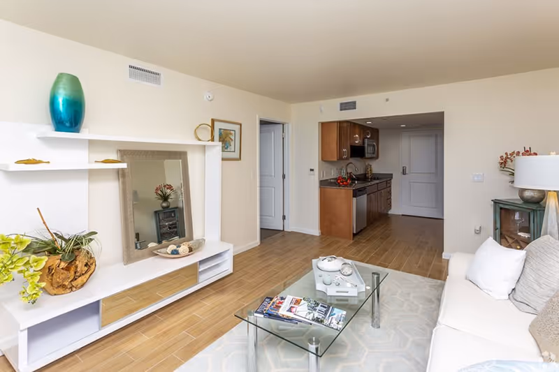 A bright and modern living room area with a white couch adorned with pillows, a glass coffee table with magazines and decorative items, a white entertainment unit with plants and a large mirror, and a small kitchen area with wooden cabinets and stainless steel appliances in the background.