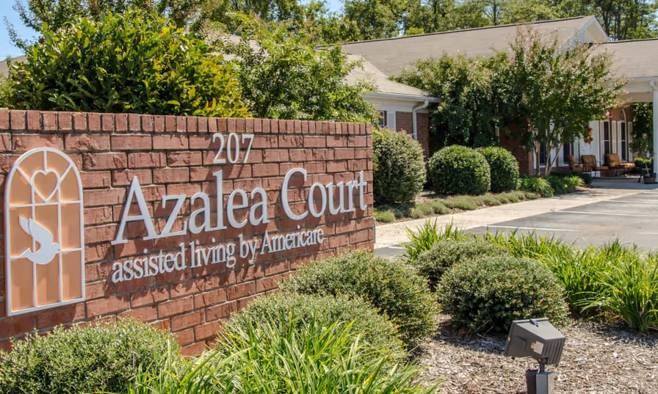 Exterior view of Azalea Court assisted living facility showing a brick sign with the facility name and logo, surrounded by green bushes and plants, with part of the building and parking area visible in the background under a clear sky.