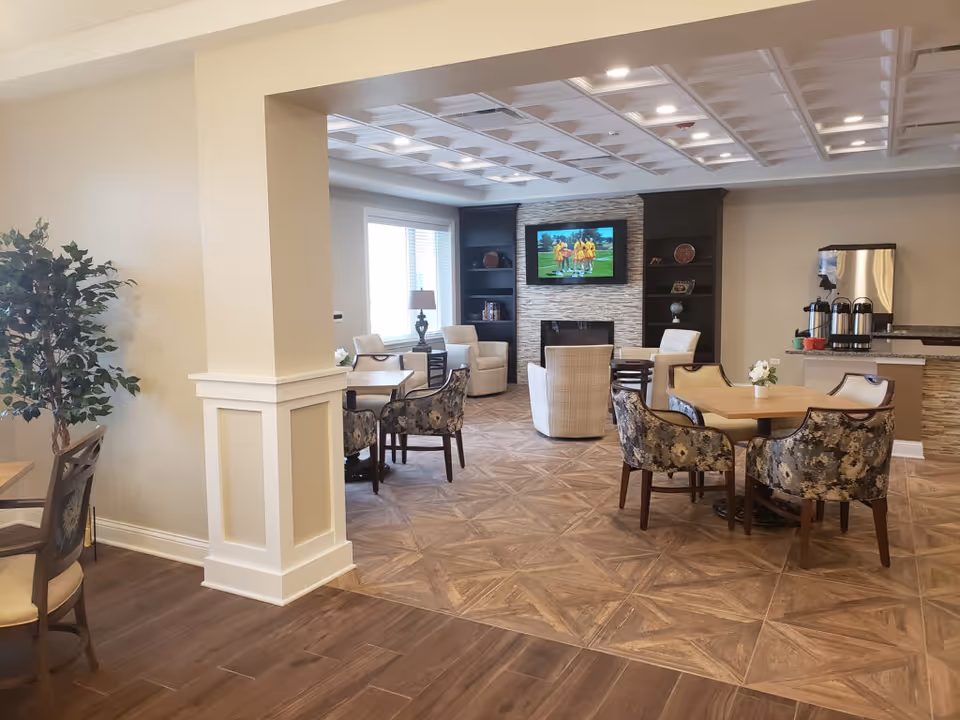 A cozy senior living common area with multiple seating arrangements including floral upholstered chairs and beige armchairs. There is a flat-screen TV mounted above a fireplace with built-in shelves on either side. A coffee station with stainless steel dispensers is visible on the right. The room has a mix of wood and tile flooring and is well-lit with natural light from a window and ceiling lights.