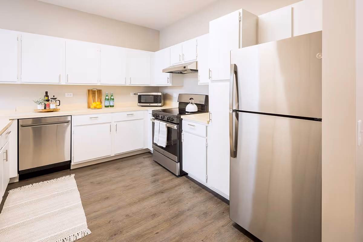 A modern kitchen with white cabinets, stainless steel refrigerator, dishwasher, and stove. The countertops are light-colored, and there is a small microwave on the counter. A white rug is placed on the wooden floor.