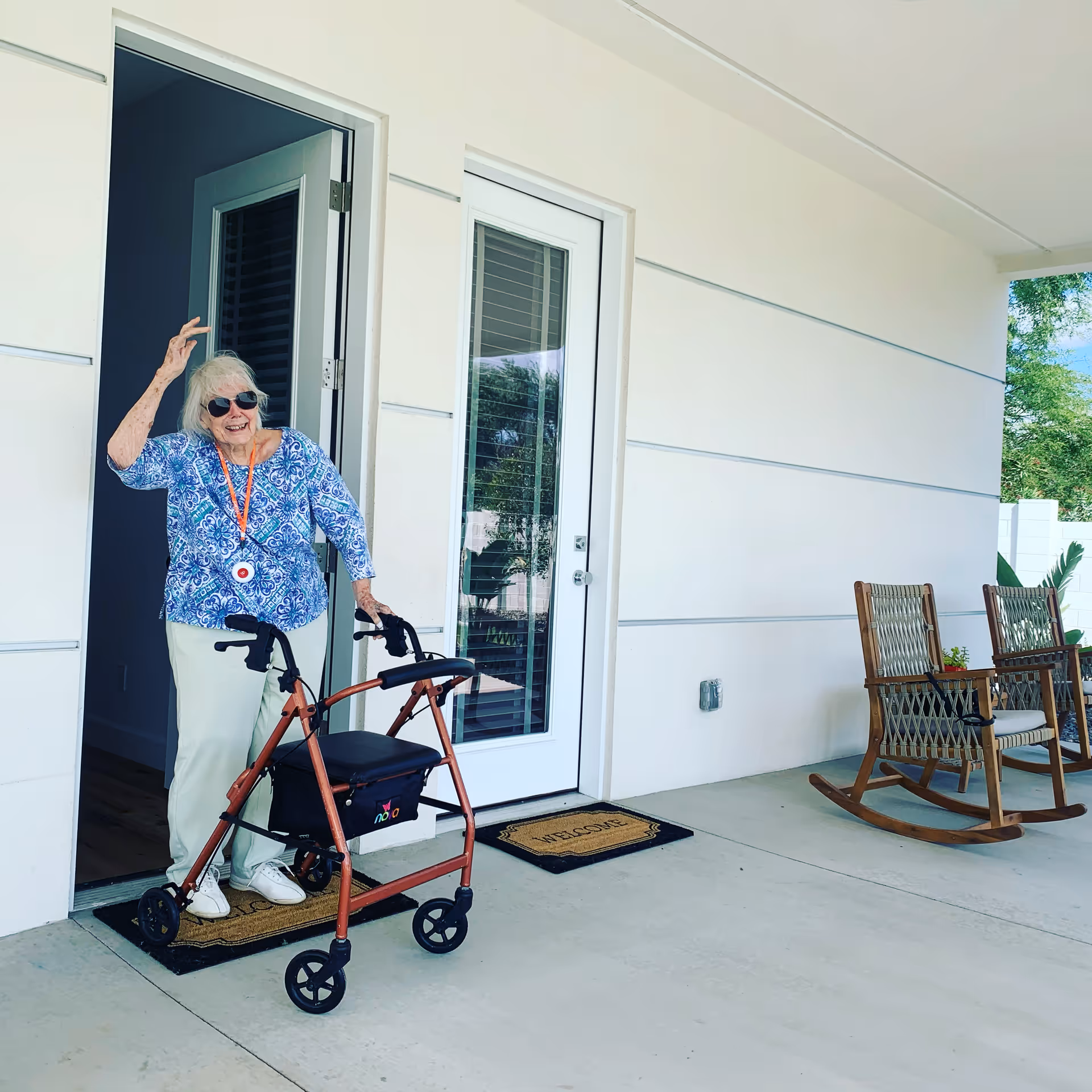 An elderly woman wearing sunglasses and a blue patterned blouse stands outside a doorway using a walker. She is smiling and waving. The porch area has two wooden rocking chairs and welcome mats in front of the doors. The setting appears to be a senior living facility.