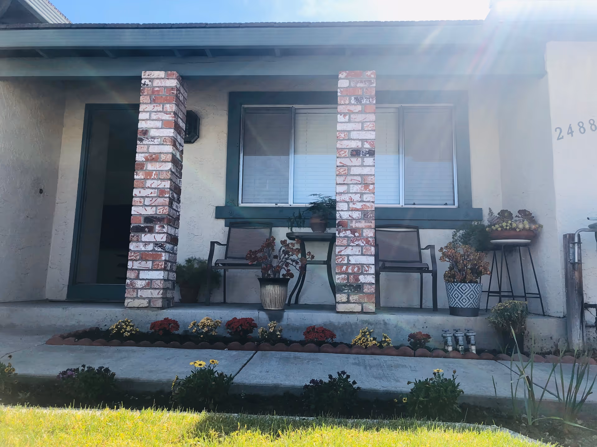 Front porch of a residential care facility with two brick columns, two chairs, a small table with a potted plant, and various potted plants and flowers arranged along the porch and garden bed. The building has a window with closed blinds and the number 2488 is partially visible on the wall.
