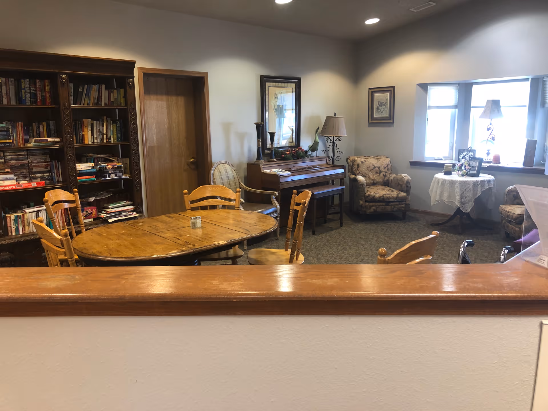 A cozy common area with a wooden oval table surrounded by chairs, a bookshelf filled with books and games, a piano with decorative items on top, two upholstered armchairs, a small round table with a lace tablecloth and framed photos, and a window letting in natural light.