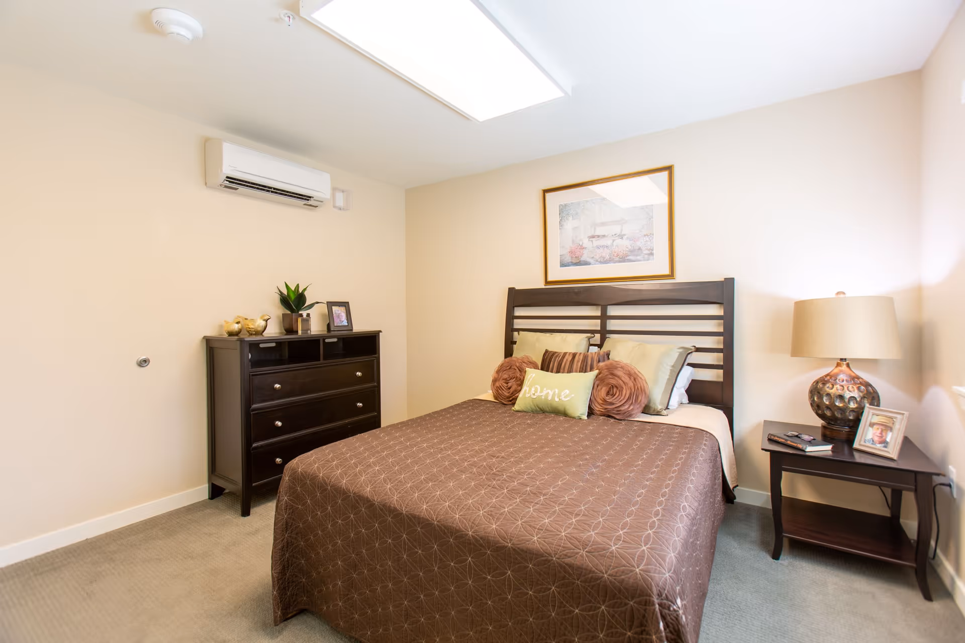 A cozy bedroom with a double bed covered in a brown patterned bedspread and several pillows, including one with the word 'home'. There is a dark wooden headboard, a matching dresser with decorative items and a framed photo, and a nightstand with a lamp, a framed photo, and a remote control. The walls are light beige, and there is a framed picture hanging above the bed. A wall-mounted air conditioning unit is visible above the dresser.