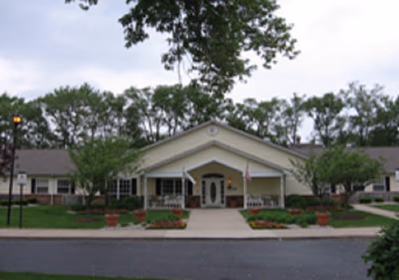 Front exterior view of a single-story building with a peaked roof entrance, surrounded by trees and landscaped flower beds, with a paved driveway and sidewalk leading to the main door.