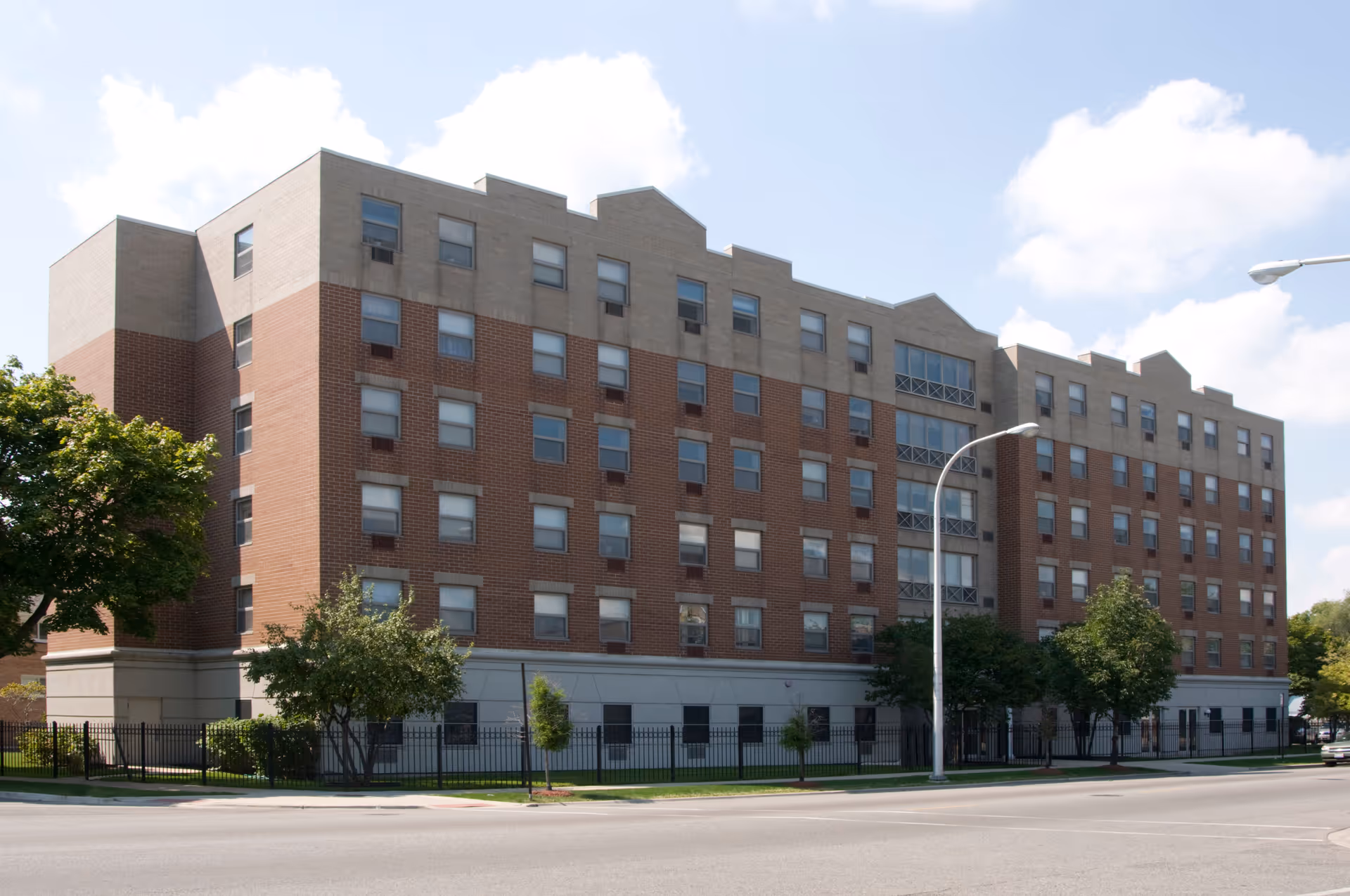 Exterior view of a multi-story senior living facility building with a combination of brick and light-colored concrete facade, multiple windows, some trees and a street lamp in front, under a partly cloudy sky.