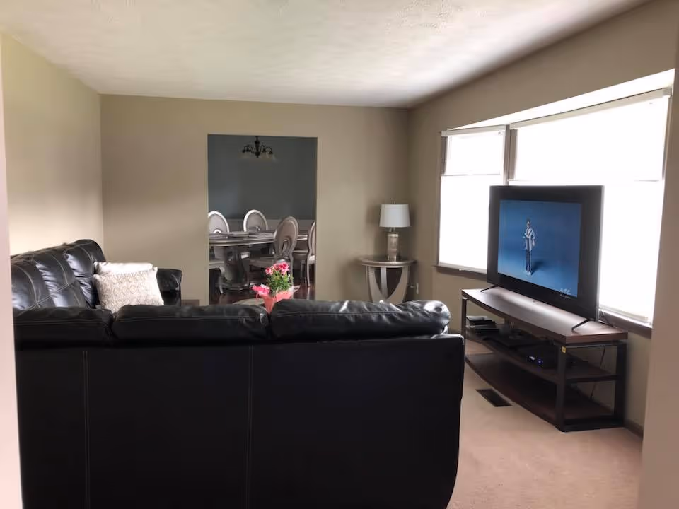 A living room with a black leather couch facing a flat-screen TV on a wooden stand. There is a small side table with a lamp next to the couch. Through an open doorway, a dining room with a table and chairs is visible. The room has beige walls and carpeted floors.