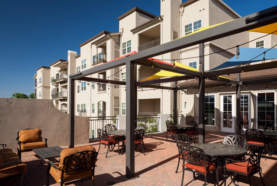 Outdoor patio area at Franklin Park Alamo Heights with metal tables and chairs featuring red cushions, shaded by colorful triangular fabric canopies and string lights, adjacent to a multi-story beige building under a clear blue sky.
