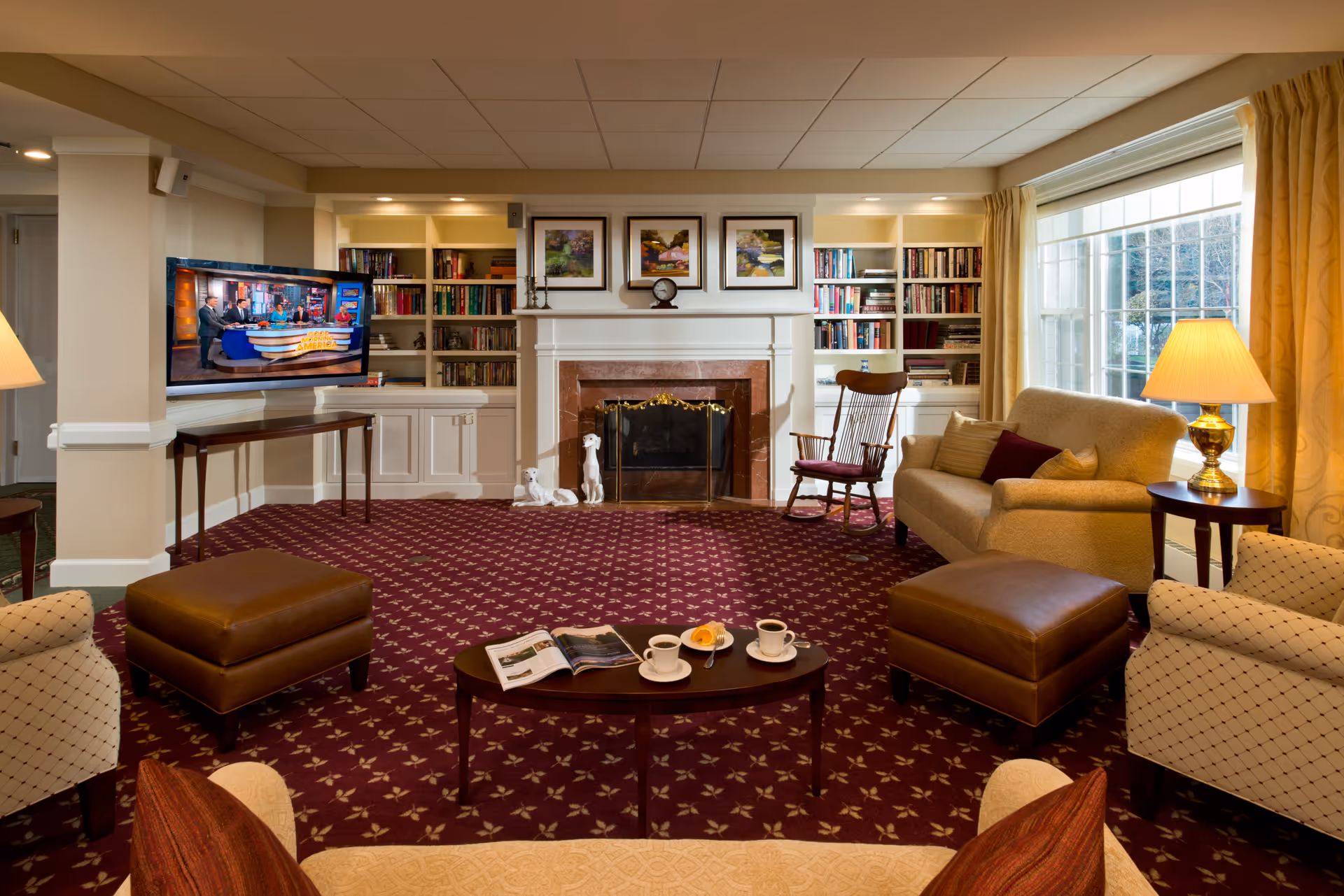 A cozy living room with patterned carpet, beige armchairs, a sofa, two brown ottomans, a wooden rocking chair, and a coffee table with cups and a magazine. There is a fireplace with a marble surround and a mantel decorated with a clock and framed pictures. Built-in bookshelves filled with books flank the fireplace. A large window with yellow curtains lets in natural light, and a table lamp is on a side table next to the sofa. A TV is mounted on a wall to the left.