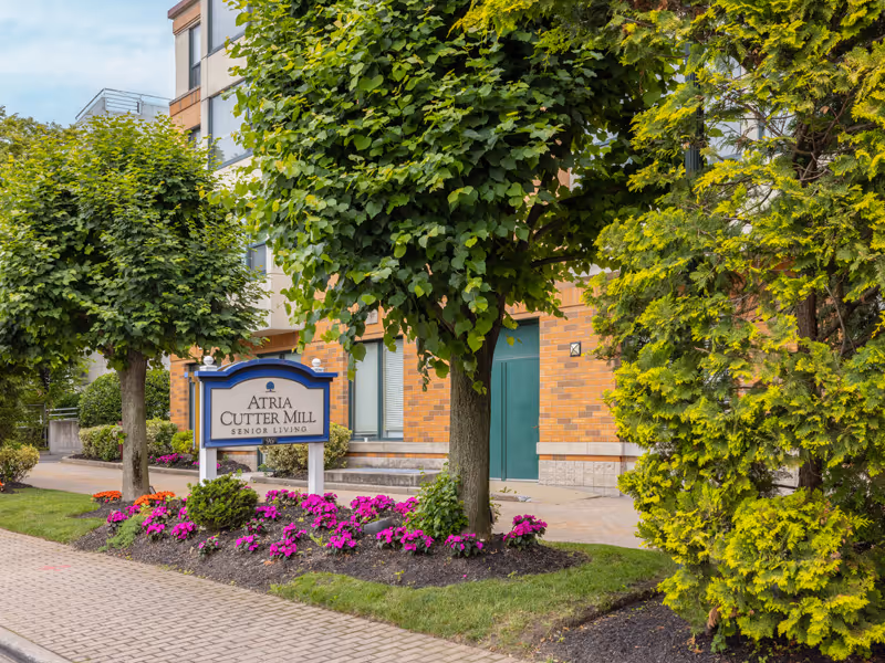 Exterior view of Atria Cutter Mill senior living facility with a sign in front surrounded by trees and flower beds, and a brick building in the background.