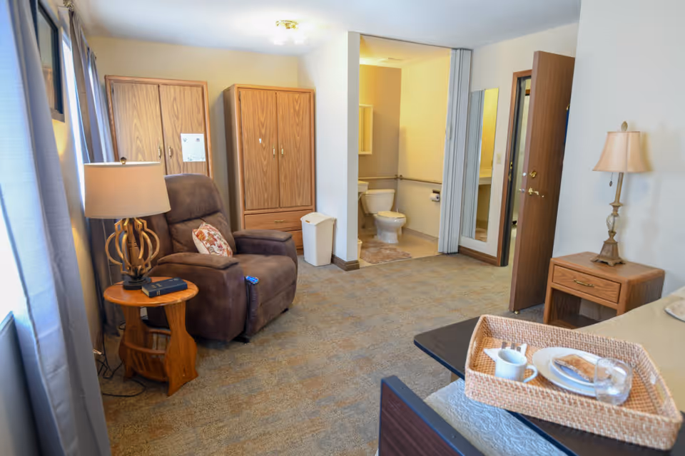A cozy senior living room with a brown recliner chair next to a wooden side table with a lamp and a book. There are two wooden wardrobes against the wall. An open doorway reveals a bathroom with a toilet and grab bars. A bed with a tray holding a cup, plate with food, and a glass of water is partially visible. The room has carpeted flooring and neutral-colored walls.