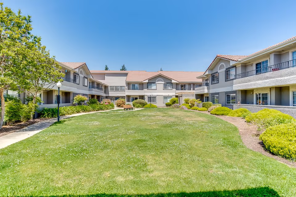 A sunny outdoor courtyard area at Fresno Senior Living featuring a well-maintained green lawn, landscaped bushes, and trees. The courtyard is surrounded by a two-story building with balconies and windows, under a clear blue sky.