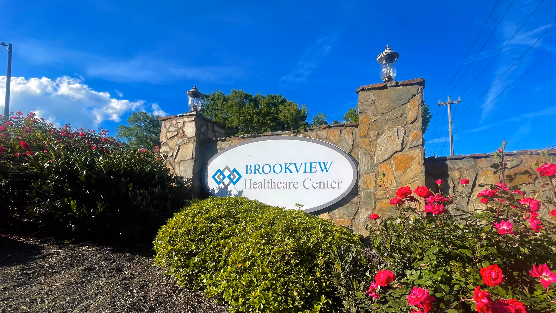 Stone entrance sign for Brookview Healthcare Center surrounded by green bushes and vibrant pink flowers under a bright blue sky with some clouds.