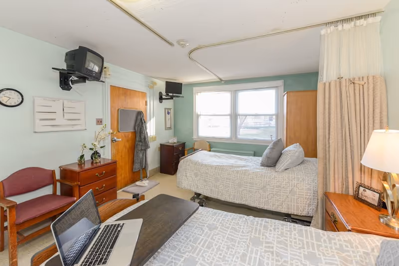 A bright shared patient bedroom with two hospital-style beds, bedside tables, a wall-mounted TV, a privacy curtain, and wooden furniture.