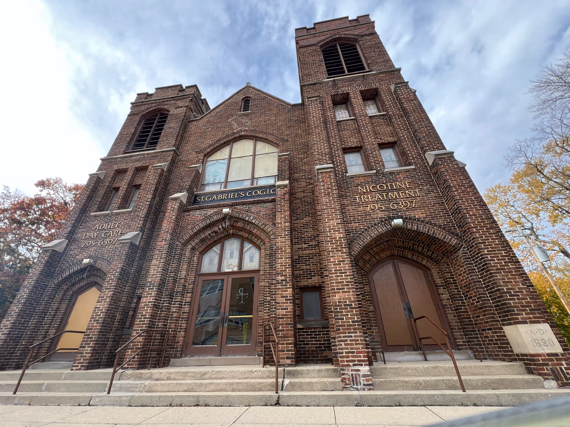Front exterior view of a large brick building with three arched doorways and two tall towers. The building has signs indicating 'Adult Day Care 795-6397' on the left tower, 'St. Gabriel's C.O.G.I.C' above the central doorway, and 'Nicotine Treatment 795-6397' on the right tower. There are steps leading up to each doorway and some autumn trees visible on the sides.