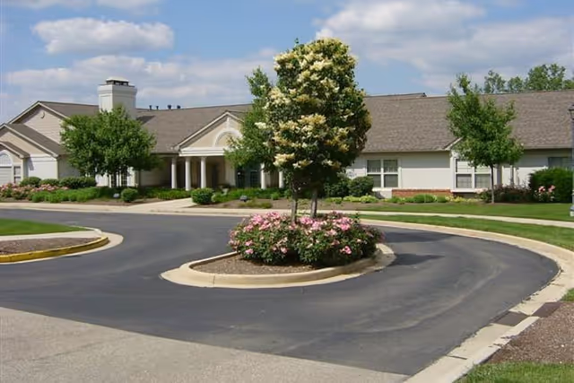 Exterior view of a single-story senior living facility building with a circular driveway and landscaped island featuring a tree and pink flowers under a partly cloudy sky.