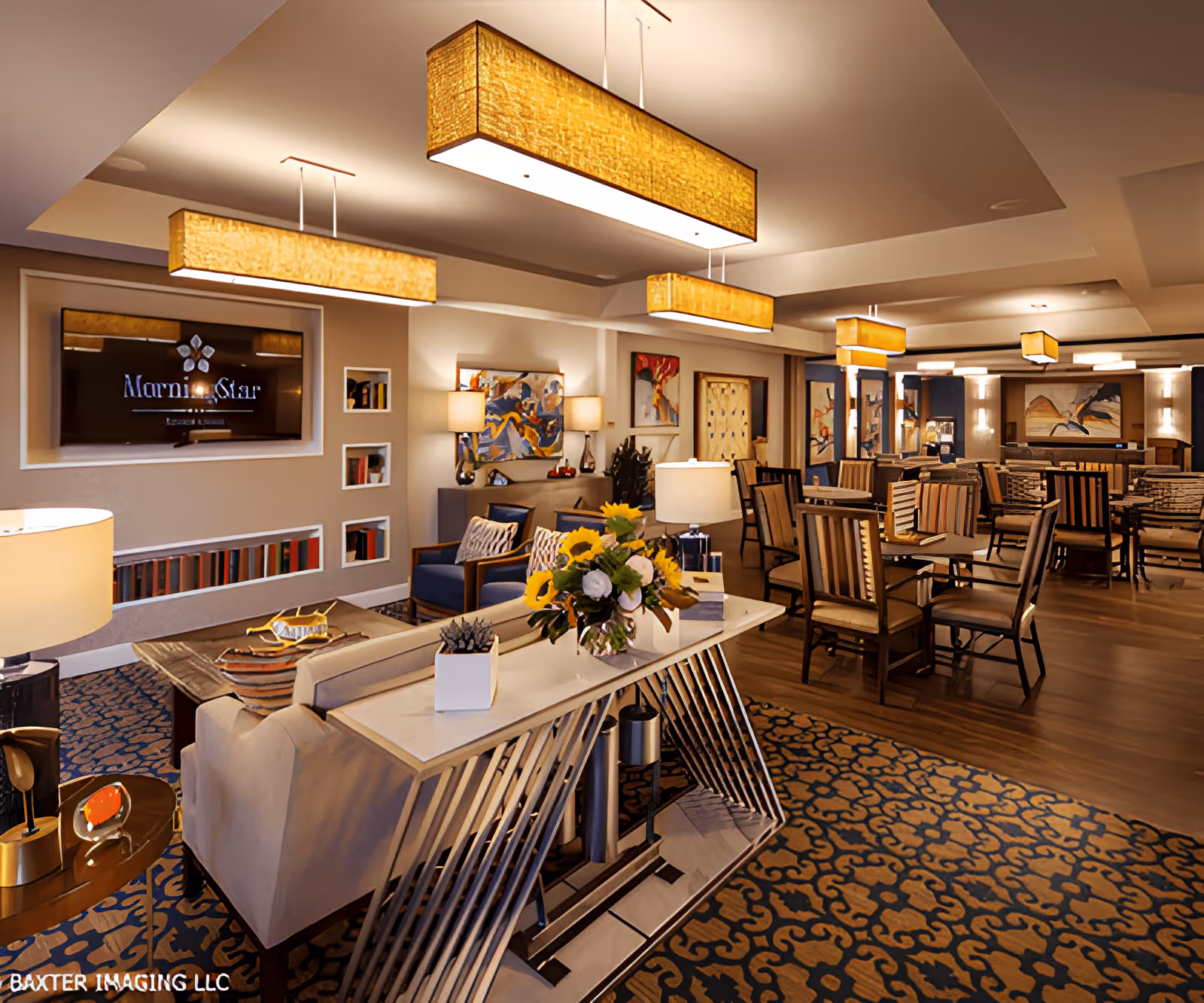 A spacious, well-lit common area in MorningStar Assisted Living & Memory Care of Rio Rancho featuring comfortable seating with sofas and armchairs, decorative lamps, a large flat-screen TV mounted on the wall, and several dining tables with chairs. The room is decorated with colorful abstract paintings, a patterned carpet, and modern rectangular ceiling lights. A console table with a flower arrangement and decorative items is positioned behind a sofa.