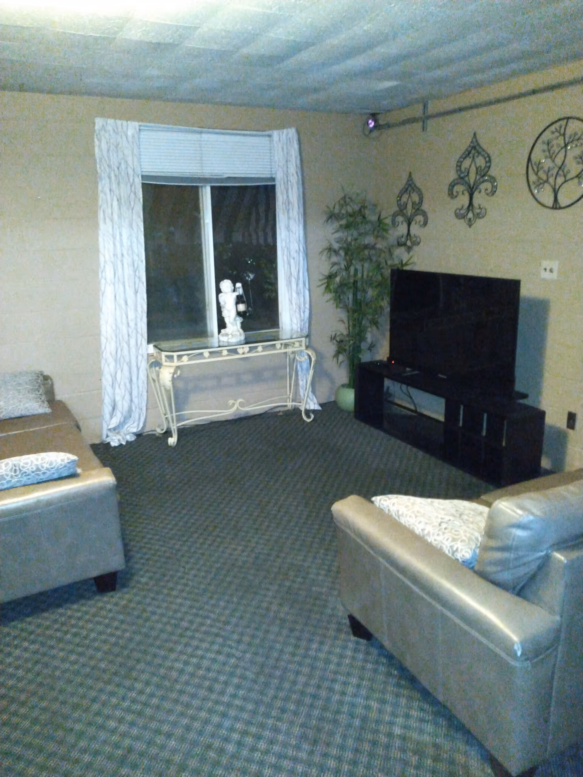 A living room with two beige leather sofas, a flat-screen TV on a black TV stand, a small decorative table with a statue in front of a window with white curtains, a potted plant in the corner, and decorative wall hangings.