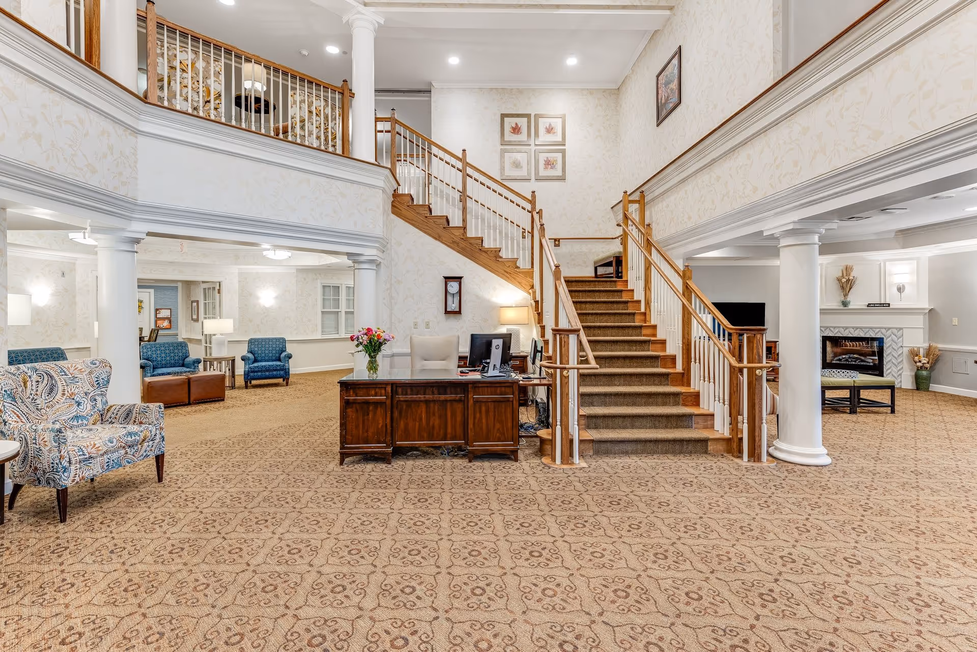 Bright senior living facility lobby with a central wooden staircase, reception desk, columns, and multiple seating areas.