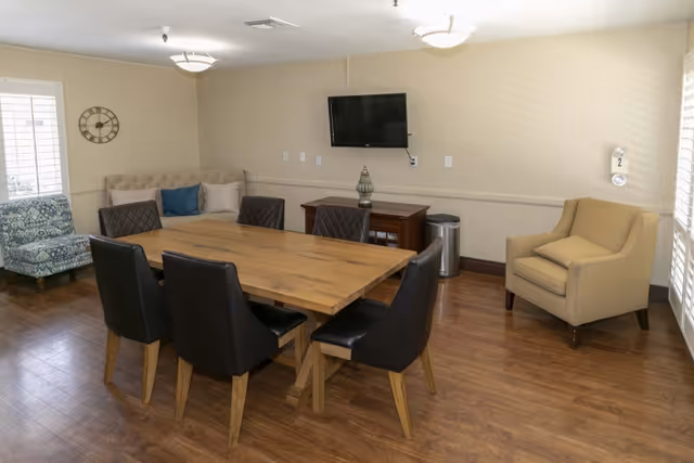A room with a wooden table surrounded by six black chairs, a beige armchair, a patterned blue armchair, and a beige tufted bench against the wall. A flat-screen TV is mounted on the wall above a wooden cabinet, and the floor is wooden. The room is well-lit with ceiling lights and natural light from windows with white shutters.