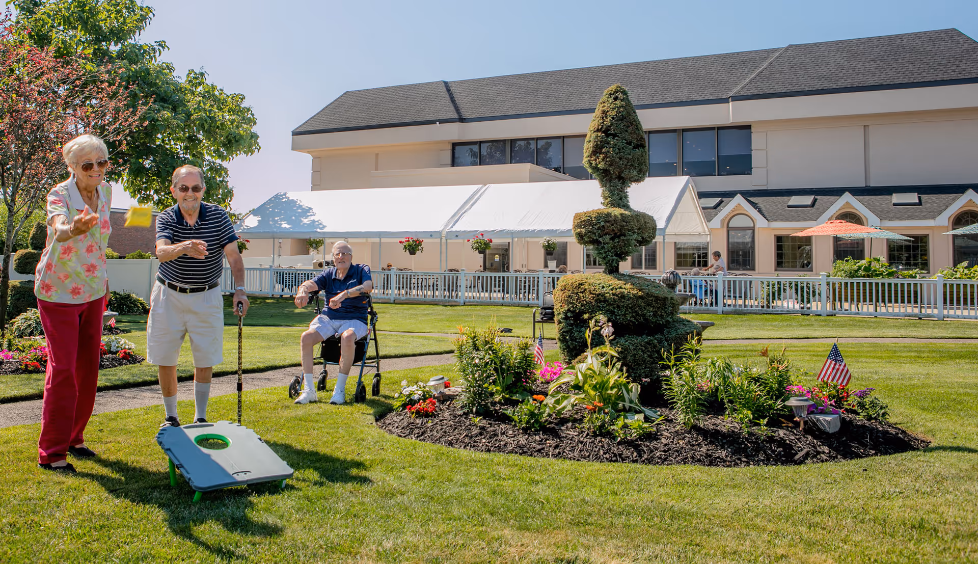 Three elderly people enjoying an outdoor game on a well-maintained lawn with a large building and white tent in the background. One woman is tossing a bean bag towards a cornhole board, while a man with a cane stands nearby and another man sits in a wheelchair watching. The garden area features a topiary and colorful flowers with small American flags.