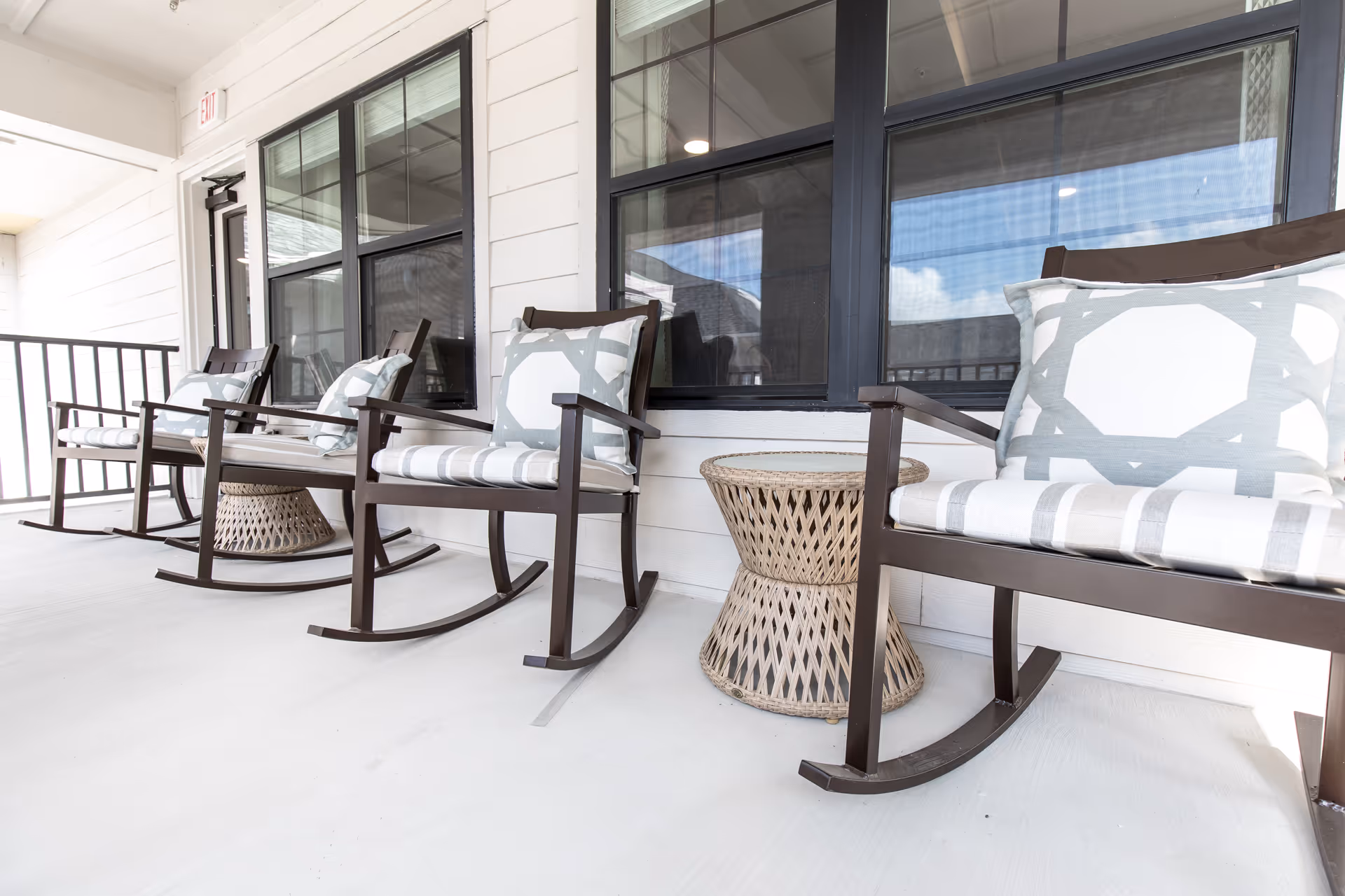 A porch area with four dark wooden rocking chairs, each with a white and gray patterned cushion, arranged in a row along a white exterior wall with large windows. Between the chairs are two woven wicker side tables. The porch floor is light-colored and there is a railing on the left side.