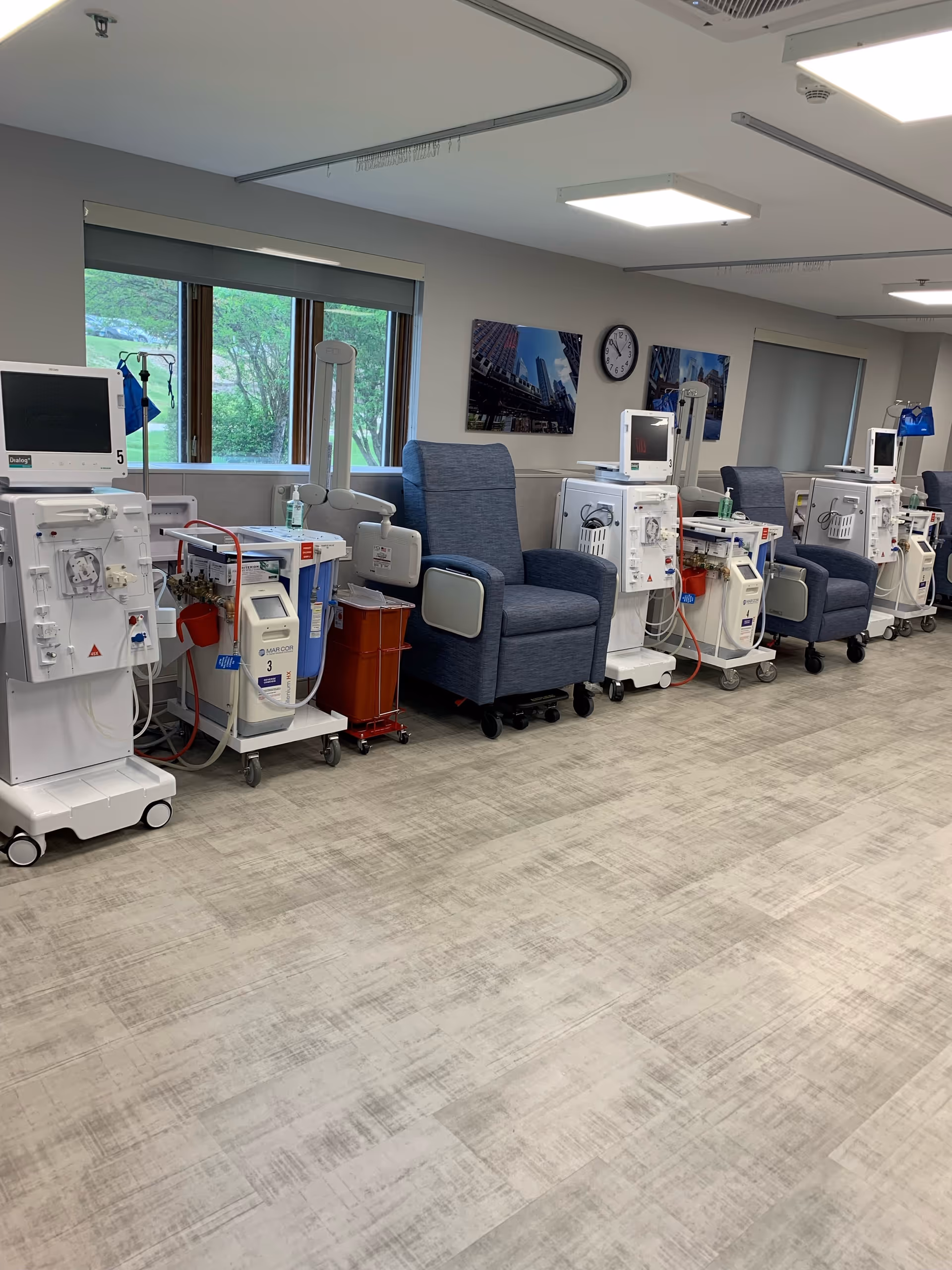 A medical treatment room with several dialysis machines lined up against the wall, each paired with a comfortable reclining chair. The room has large windows with a view of greenery outside, light-colored flooring, and framed pictures on the wall.