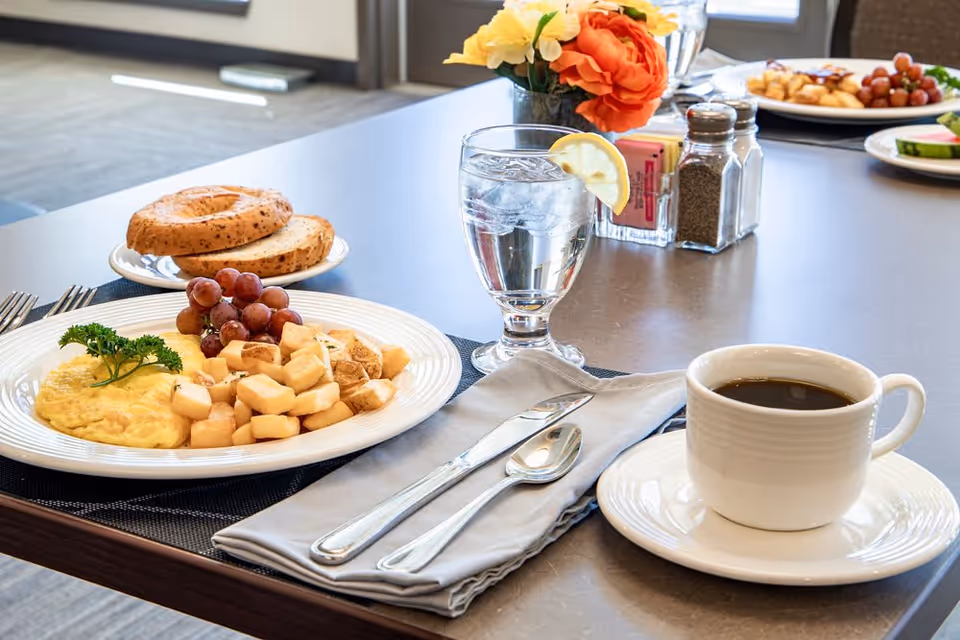 A breakfast meal on a table including an omelette garnished with parsley, diced potatoes, red grapes, a plate with two bagel halves, a glass of water with a lemon slice, and a cup of black coffee. The table also has a napkin with a fork and spoon, salt and pepper shakers, a small container with sugar packets, and a vase with orange and yellow flowers.