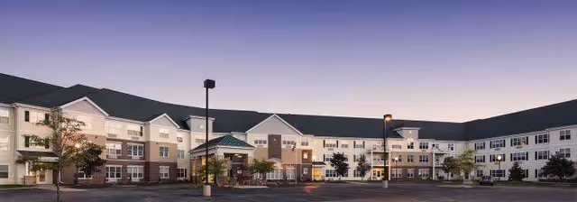 Wide exterior view of a large, multi-story senior living facility building at dusk with well-lit windows and a spacious parking lot in front.