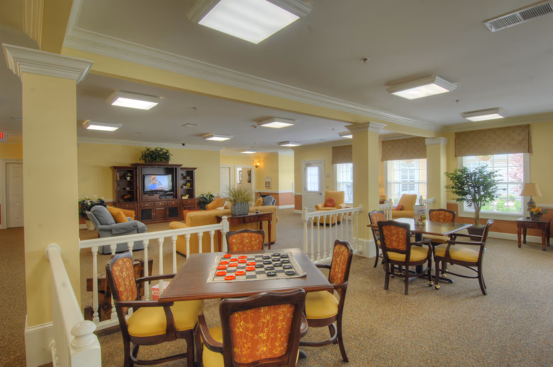 A spacious and well-lit common area in a senior living facility with multiple seating arrangements. In the foreground, there are tables with chairs, one table featuring a checkers game set up. In the background, there is a living room area with sofas, armchairs, a wooden entertainment center with a television, and large windows letting in natural light. The walls are painted a soft yellow, and the carpet is beige. Potted plants and decorative items add a homey touch.