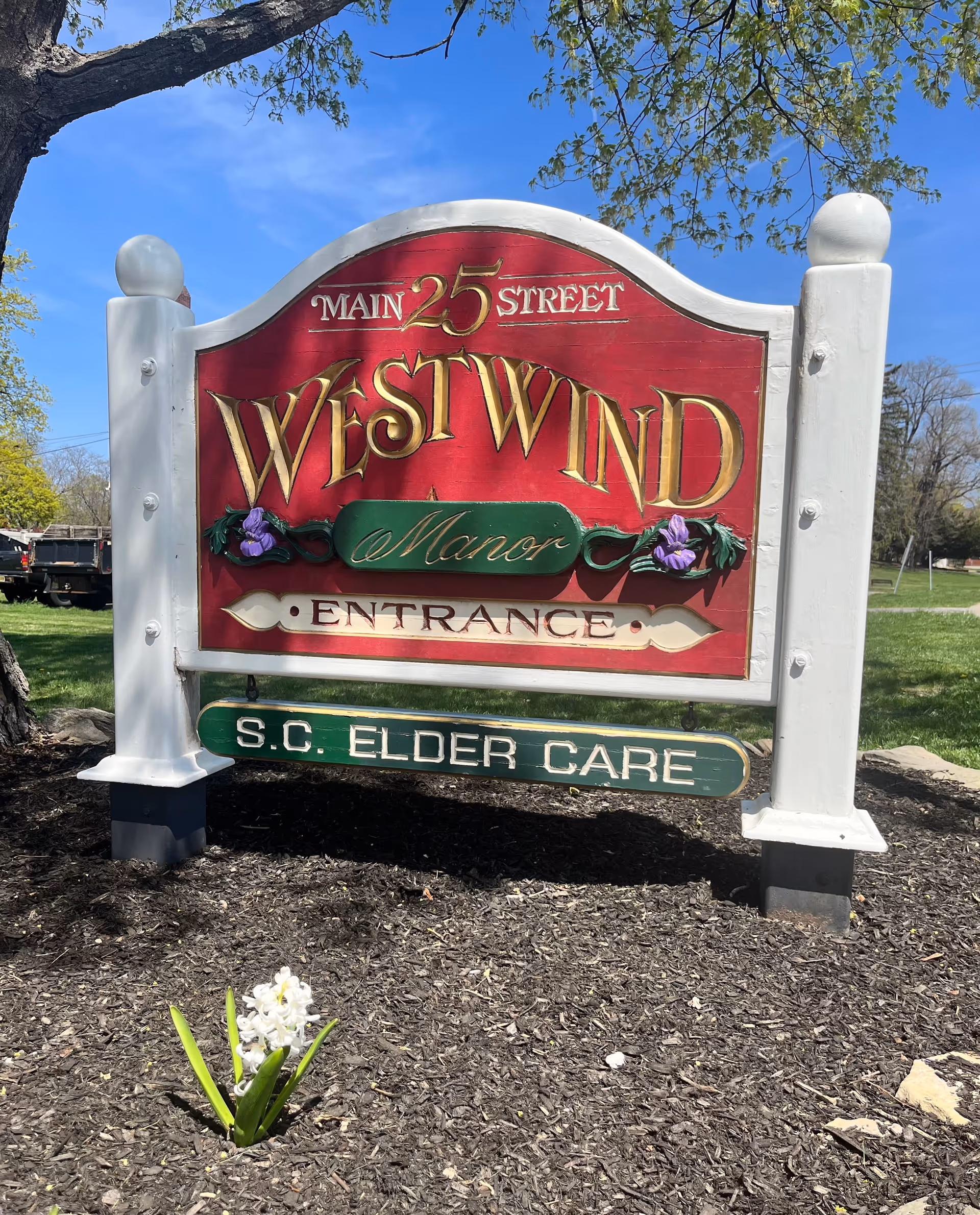 A decorative outdoor sign for Westwind Manor, located at 25 Main Street, with the words 'Entrance' and 'S.C. Elder Care' displayed. The sign is red with gold and green accents, mounted on white posts, with a small white flower planted in front and a tree and grassy area in the background under a clear blue sky.