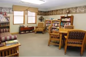 A cozy living room area with beige carpet, two windows with blinds and valances, a striped sofa, a wooden bookshelf filled with books, a wooden table with four cushioned chairs, and two armchairs with patterned upholstery. The walls have a decorative border near the ceiling.