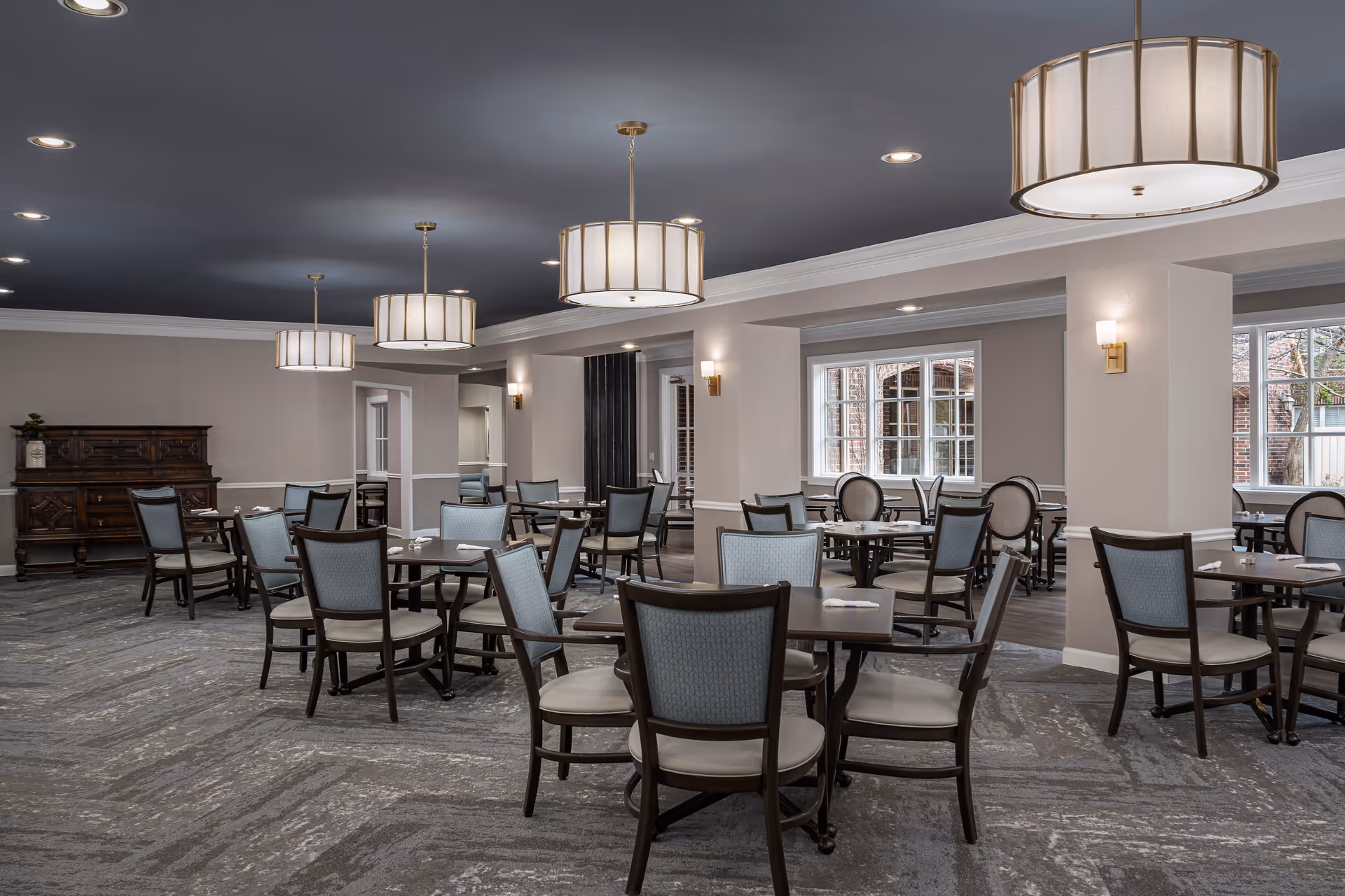 A spacious dining room in an assisted living facility with multiple round tables and chairs arranged neatly. The room features large windows letting in natural light, modern pendant lights hanging from a dark ceiling, and a carpeted floor with a patterned design. There is a wooden sideboard against one wall.