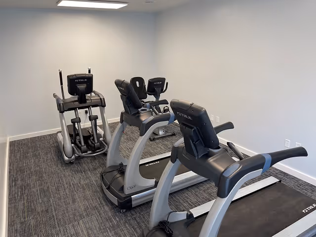 A small fitness room with two treadmills and two stationary bikes against white walls and gray carpeted floor.