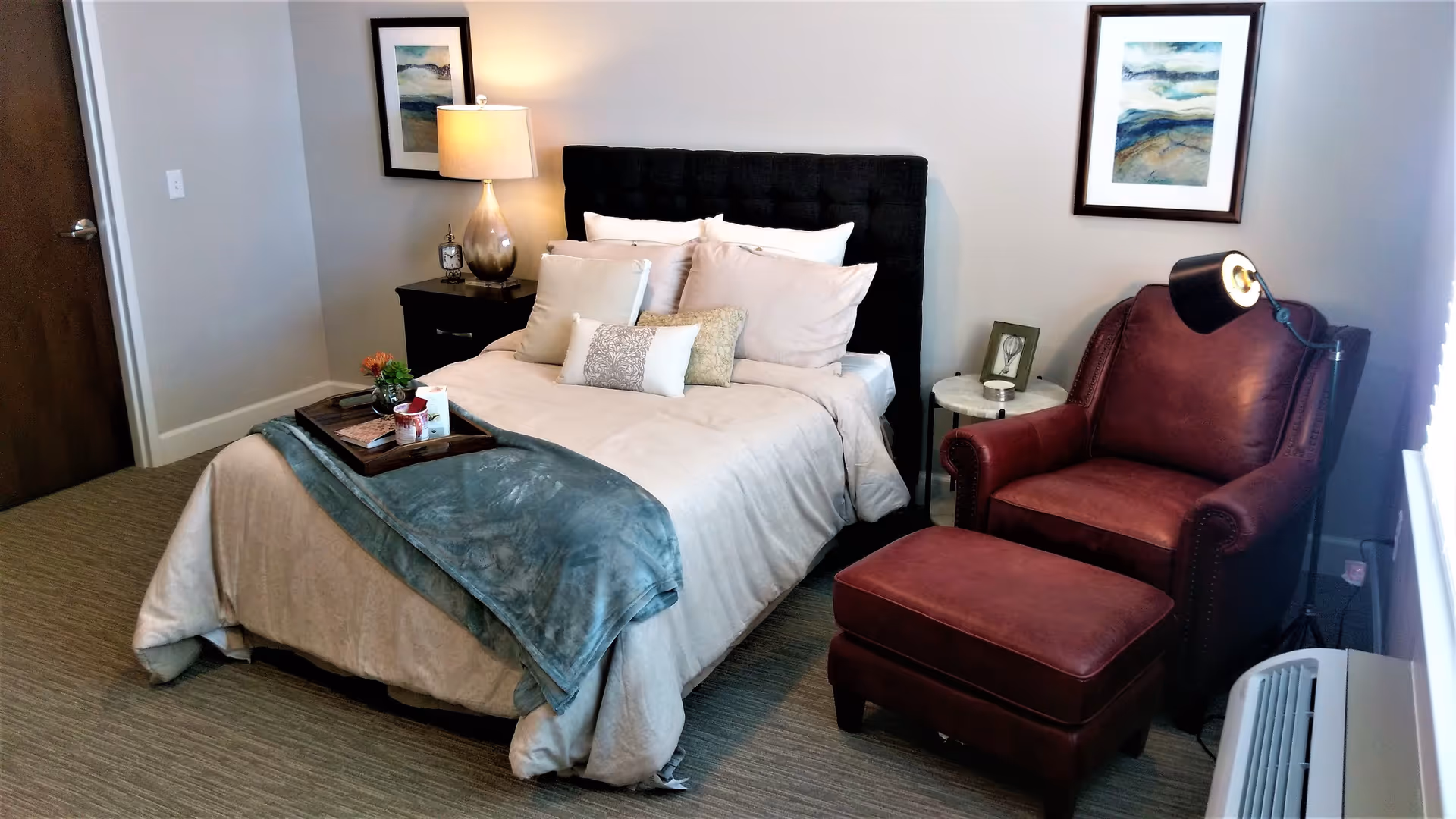 A cozy bedroom with a neatly made bed featuring multiple pillows and a blue throw blanket. There is a dark upholstered headboard, a wooden nightstand with a lamp and clock, a red leather armchair with a matching ottoman, a small round side table with a framed picture and candle, and two framed landscape paintings on the walls.