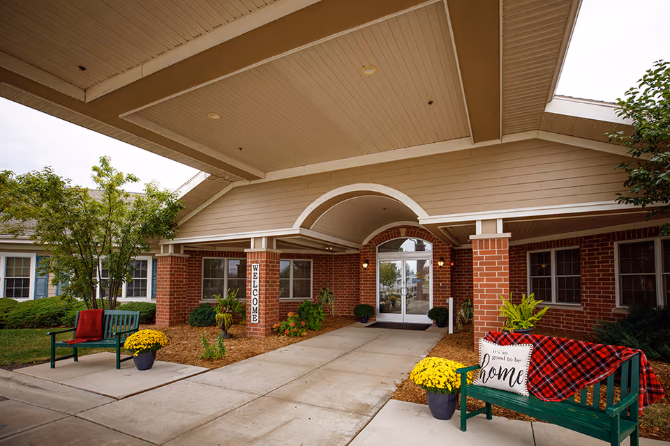 Entrance of Parkside Manor Assisted Living and Memory Care with a covered walkway, brick pillars, and glass double doors. There are two green benches on either side of the walkway, one with a red blanket and the other with a red plaid blanket and a pillow that says 'It's so good to be home'. Yellow potted flowers and green plants decorate the area.
