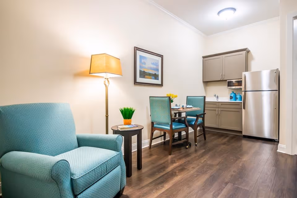 A cozy interior space featuring a light blue upholstered armchair next to a small round wooden side table with a potted plant and a book. A floor lamp with a beige shade stands behind the table. In the background, there is a small dining table with two teal cushioned chairs, set with plates and cups. Above the table hangs a framed landscape painting. To the right, a compact kitchenette includes taupe cabinets, a stainless steel refrigerator, a microwave, and a small sink. The room has wood flooring and cream-colored walls.