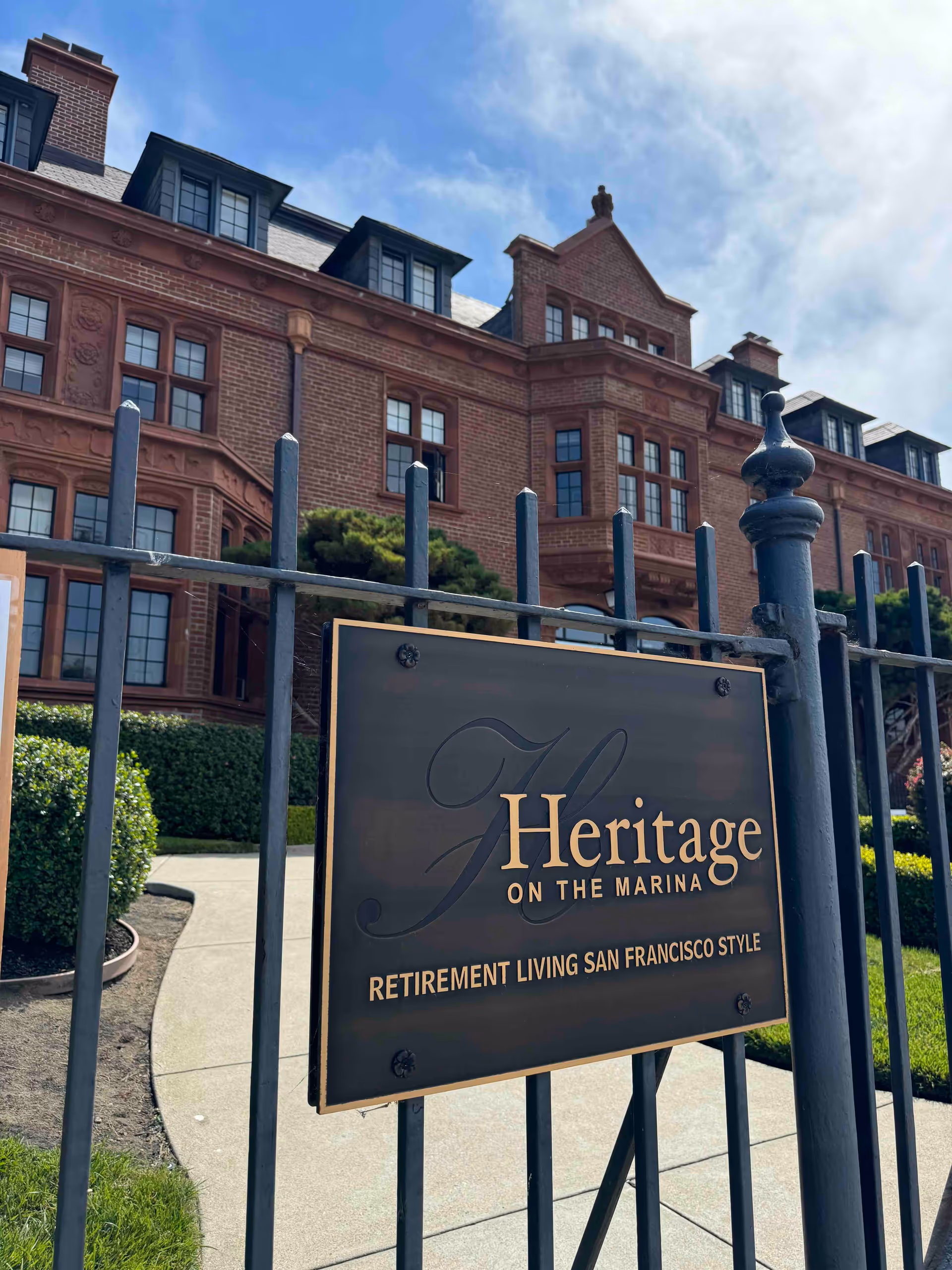 Metal sign reading "Heritage on the Marina" mounted on a wrought-iron fence in front of a large red-brick building.