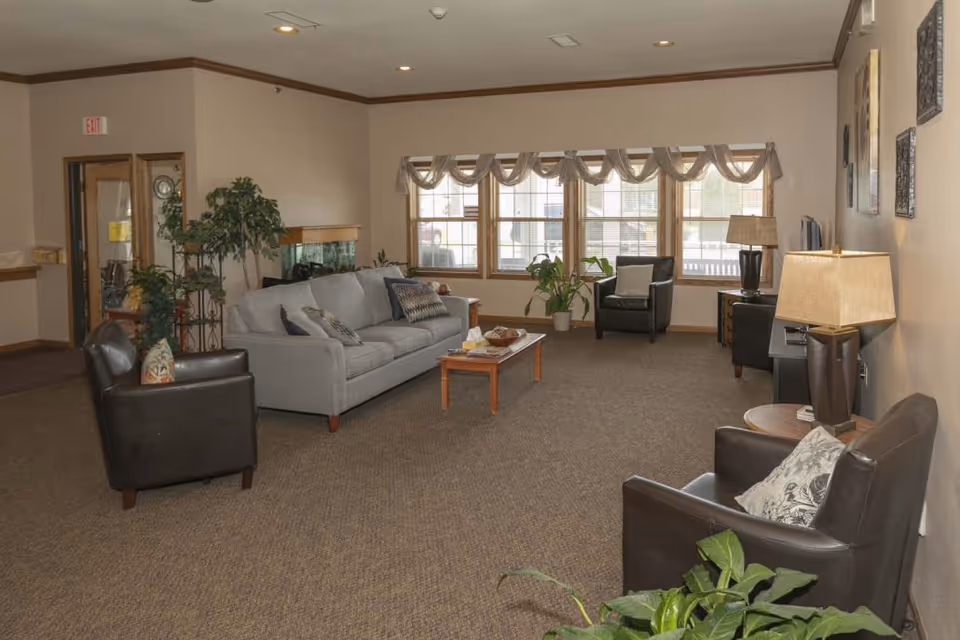 A spacious living room in an assisted living facility with a gray sofa, three dark leather armchairs, a wooden coffee table, several potted plants, a large window with valance curtains, and lamps providing warm lighting.